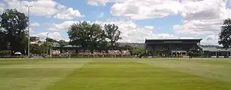 A cricket ground with two stands and several trees visible.