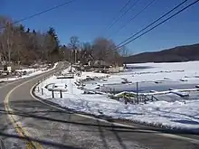 A road straddling the bank of a frozen lake. The surrounding ground is covered by thin layer of snow. Powerlines are visible overhead