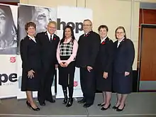 A photograph of six standing, smiling people, five of whom are wearing black uniforms with the sixth wearing a pink dress shirt