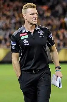 A male athlete with light hair wearing a polo shirt and pants walks on the grass surface of the playing arena.