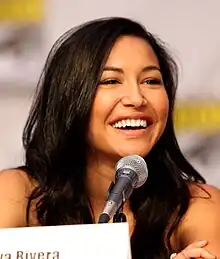 A young woman in a black fitted suit smiling and holding an award statuette in each hand.