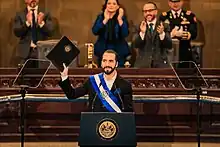 Nayib Bukele standing at a podium and holding up a legislative proposal in his right hand