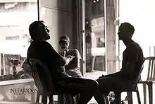 A black-and-white photograph of three men sitting on chairs facing each other and wearing shorts and T-shirts all next to another empty chair