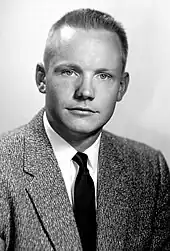 A black-and-white photo of a young man with light skin and pale irises. His mid-colored hair is cut short. He is looking at the camera. He is wearing a barleycorn sport coat, a white shirt and a dark necktie.