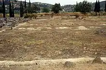 Image of the remains of the first oikos taken in 1991. The west wall is in the foreground and the limestone walls are very short and damaged. Only the rectangular walls remain around the soil and grass, there are also hills and trees in the background.