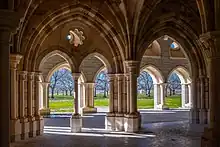 Multiple interior and exterior stone arches frame a view looking out from a chapter house, revealing a view of orchard trees beyond an expanse of bare ground.