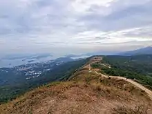 Hiking trail in foreground, winding down to a long, narrow plateau in the midground; small coastal town, inland sea and outlying islands in background