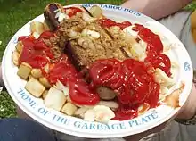 Picture of Garbage Plate. Plate says "Nick Tahou Hots" and "Home of the Garbage Plate" Plate includes two hots, potatoes, the rest is covered in catsup.