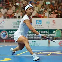 A young female squash player in white, stretching out to get her racket under a low ball, on a blue court with yellow markings, in front of a large, out-of-focus crowd behind glass.