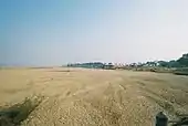 A daytime view of a desiccated, scappled expanse that wholly fills the fore- and middle ground and arcs away to the horizon at left. Of clouds in the sky there are none whatever. At extreme right in the middle distance, a thin bank of trees sits on a slightly higher level, reaching to the middle and rightward horizon; people and livestock mill about before them. Several pink buildings, perhaps a village, sit similarly elevated in front of the trees at middle-right, beyond the dominating dusty expanse.