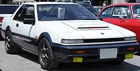 Front-three-quarter view of a two-door car with pop-up headlights, a hood scoop, and door mirrors