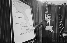 A room with a curtain and an American flag in the background. A man in a suit points to Cambodia on a large standing map of Southeast Asia.