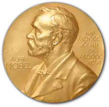 A golden medallion with an embossed image of a bearded man facing left in profile. To the left of the man is the text "ALFR•" then "NOBEL", and on the right, the text (smaller) "NAT•" then "MDCCCXXXIII" above, followed by (smaller) "OB•" then "MDCCCXCVI" below.