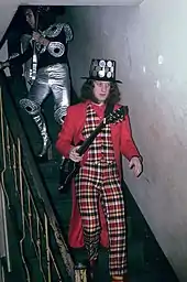 A man walks down a flight of stairs. He holds a guitar and wears a black hat studded with round metal mirrors. His waistcoat and trousers are tartan. His coat and shirt are plain red. His socks are striped.