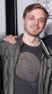 A white man with blue eyes and blonde hair styled in an undercut. He is wearing a gray T-shirt and jacket, and a hand is resting on his left shoulder.