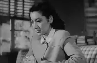 Noriko, wearing the same dress as in the Noh sequence, is shown seated on a sofa from a slightly lower angle; a window and some books are dimly visible in the background.