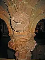 The top of a column and base of two arches. In the spandrel is a carving of an otter-like creature; the capital is decorated with a carved head and foliage