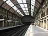 A railway station with side platforms either side of two tracks that disappear into darkness under a painted steel bridge like structure topped with a brick wall, covered by a partially glazed barrel roof.