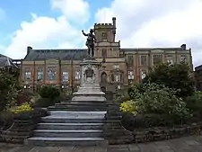 Photograph of a Gothic Revival building in grey slate, with walls of light brown and a pitched roof bearing algae. It has a wide chimney on each side, five large multi-segment windows on each storey on each section. The left and right sections have two and three storeys respectively, separated by a central protrusion with five storeys. The central section has two-storey bays window flanking an arched, elevated wooden door, above an outdoor double staircase with a simpler wooden door in the front. Above each bay window is a smaller window. Above these is a narrower section with three large multi-segment windows, above which is a yet narrower section forming a square tower and the highest storey. The tower is crenellated with one large multi-segment window, a flagpole with no flag in the centre, and a crenellated stone structure projecting from the right corner like a crow's nest, which could be used as a lookout. The building is fronted by a lawn between an embankment and a planted area. Steps down from the central part lead to a war memorial: a bronze statue of a First World War uniformed British soldier looking back and pointing up towards the left, atop a white stone plinth with a relief of Nottingham's heraldic achievement, an inscription on a plaque and a relief of the lozenge of Dame Agnes Mellers. The war memorial has stone steps on all four sides separating an outer circle of shrubbery. Below is a paved stone floor.