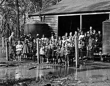 A group of schoolchildren and their teacher standing in front of a small school building