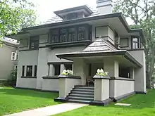 Recent exterior view from the northeast corner.  In the foreground is the front porch with broad steps and planting piers which hides the front door in shadow.  The rest of the house rises behind.  A flat yard of turf grass extends up to the base of the house with no other landscaping.