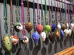 Colourful four-hole English pendant ocarinas hanging from a rail by elaborately-knotted 2- and 3-colour lanyards. Three are shaped like squat owls; several have low-relief or painted patterns.