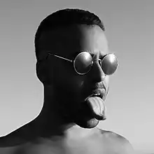 A black-and-white picture of Twin Shadow. He wears teashades and he sticks his tongue out.