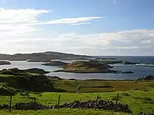 A view from a low eminence looking towards a fence and a tumble-down stone wall beyond which is a strip of uncultivated land  and the sea shore. A variety of  hummocky green and brown islands hug the coast as waves line their rocky shores under a blue sky.