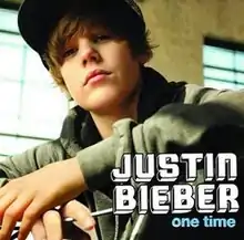 A 13-year old Justin Bieber stands in front of a building with block windows, while on the handlebars of a bicycle and wearing a jacket and fitted hat.