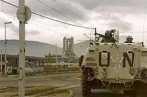 A dilapidated city block, with a street crossing in front of it from left to right. The structures are in ruin, and a multi-storied building in the center is partially collapsed. On the right, a military tank bearing the letters "UN" and carrying two visible soldiers, prepares to cross railroad tracks. In the background are large hills and a grey cloudy sky.