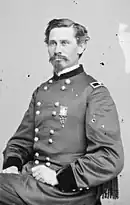 Black and white photo shows a young man seated. He has a moustache and goatee and wears a dark military uniform with two rows of buttons with the shoulder tabs of a brigadier general (one star).