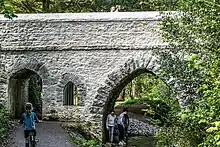 A bridge in St. Anne's Park. It is grey and with Arches of three sizes. There are trees in the background and a stream on the right side of the picture. A child is riding a bicycle, and three people are under one arch.