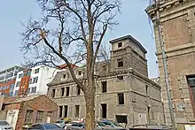 A tan building similar to the main house with a tower on the right. In front of it is a large bare tree. To the lower right is a small dilapidated brick building with a gabled roof.