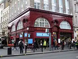 A corner view showing both elevations of a red glazed terracotta building. The first storey above ground features two wide storey height semi-circular windows on each elevation above which is a dentil cornice. Below the windows, the station name, "Oxford Circus", is displayed in gold lettering moulded into the terracotta panels. Above the first two storeys the building has been extended as offices with an ornate Portland stone façade.