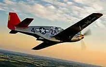 A single nosed-propeller plane is in mid flight above land.  The red nose faces right and the underside of the plane is slightly in view.  The plane has black propellers, grey base paint, and black letters reading A42.  The 4 and 2 are separated by a United States roundel in black with a central white star.  The roundel is also visible on the tops of the planes wings.  The full length of the right side of the plane is visible.