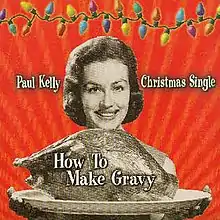 A sepia coloured image of a woman presenting a platter containing a large roast bird (possibly a turkey). Her smiling face is visible beyond the bird, her fingers appear below the platter. The background is red. A strand of coloured Christmas lights stretches across the top of the image. The artist's name is located to the left of the woman's head and the words, Christmas Single, are to her right. The title of the single is written over the image of the roast bird.