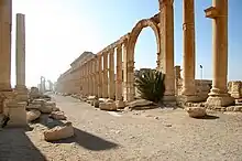 A crumbled array of stone pillars stand around an archway, lined along a dusty avenue.