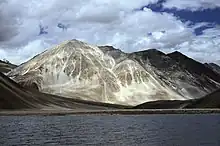 Daytime view of a large body of water standing before a prominent peak, which communicates with several others partly out of view and behind. A gravel beach at the far end of the lake gives way to steep slopes leading up to the peaks; The mountains lack trees. Patchy snowcover defines their recesses, and whitish vein-like streaks extend up from the base of the largest.