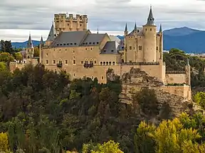 A castle high on a rocky peninsula above a plain. It is dominated by a tall rectangular tower rising above a main building with steep slate roof. The walls are pink, and covered with a sculptural pattern. There is a variety of turrets and details.