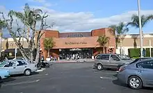 A view of a single-story brown brick building from a parking lot in front, with palm trees on either side of the frame. In the center is a red brick entrance pavilion with "Panorama Mall" written on it in stylized metal letters