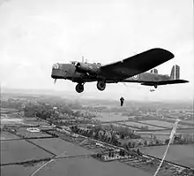 Man hangs suspended from aircraft flying over tree lined fields