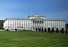 A large, symmetrical white building with classical-style columns front and centre sits uphill of the viewer, behind a well-tended lawn.