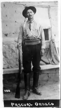 A black-and-white photograph of a man in a cowboy hat, a cartridge belt around his waist and a rifle by his foot, looking straight at the camera