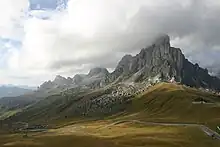 Cloud-covered mountains sit as the backdrop to a green valley below, with a serpentine road running through it.