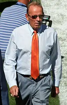 Candid photograph of Bowlen walking on a football sideline wearing blue-grey slacks, a light blue shirt with a red tie and sunglasses.