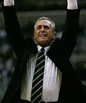 A man, wearing a jacket with a white shirt and tie, is raising his hands in the air during a basketball game.