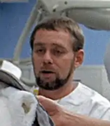 A man with short brown hair, beard and mustache wearing a white top looks to the left and down; he appears to be in a hospital room