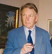 Colour photograph of Paul Britten Austin holding a wineglass in front of an oil painting