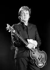 A serious, standing man holding his guitar with his left hand and placing his other hand on his left arm