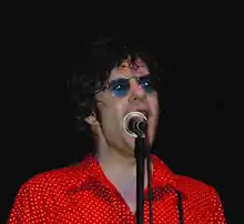 Shoulder high portrait wearing red polka dot shirt and blue sunglasses, sweating, singing into microphone which partly hides his face.